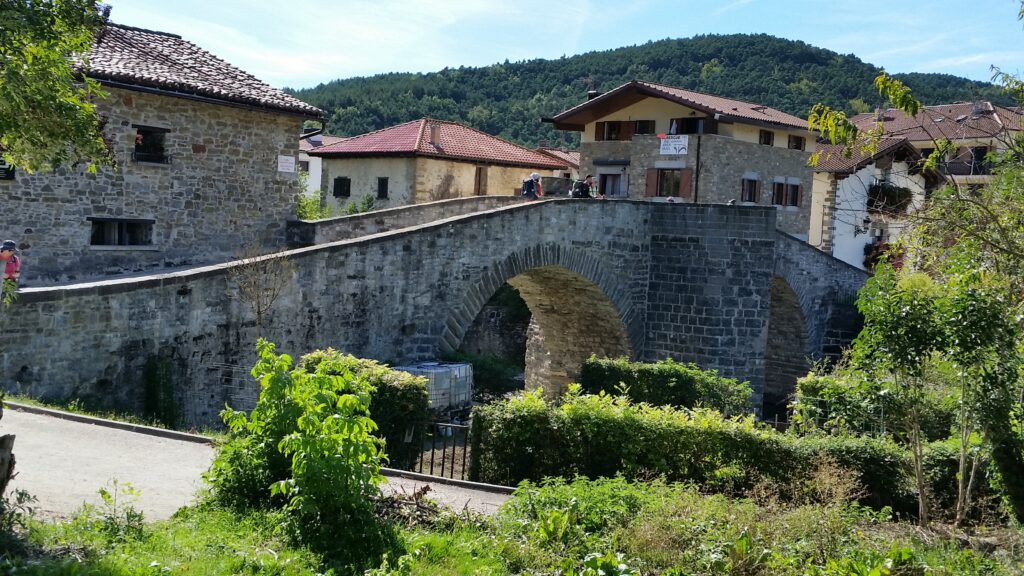 A picturesque stone bridge spans a small river in a quaint European village, nestled amidst lush greenery and rolling hills under a clear blue sky.